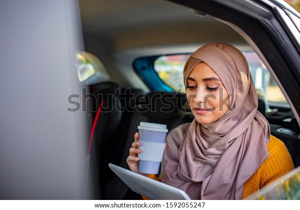 Busy young arabian woman with hijab on head sitting\
in taxi cab, typing on laptop keypad and talking on smartphone.\
Portrait of a young, attractive Muslim woman wearing a hijab\
commuting in a car.