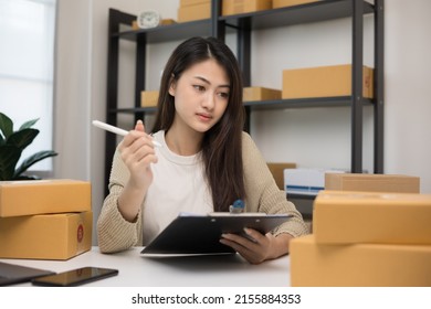 Busy Young Asian Entrepreneur Woman Checking Product On Stock Take Note With Clipboard. Business Woman Working SME Online At Home. Small Business Owner Start Up Freelance Concept.