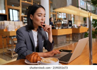 Busy Young Asian Professional Business Woman Wearing Suit Using Laptop Computer Sitting In Cafe. Chinese Lady Manager Remote Working Online Thinking On Online Marketing Operations Or Elearning.