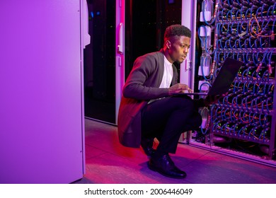 Busy Young Black Datacenter Engineer Crouching In Server Room With Neon Illumination And Checking Network Settings On Laptop