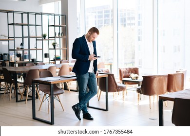 Ein beschäftigter junger Geschäftsmann mit Kleidung für Mode ist das Handy in einem modernen Bürozimmer in der Nähe eines Holzschreibens auf dem Hintergrund eines großen Fensters. Konzept der Büroarbeit.