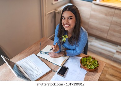 Busy Young Business Woman Eating A Healthy Lunch While Working At Home. A Working Woman Eating Lunch At Her Desk. Happy Business Woman Eating A Salad From A Bowl While She Works 