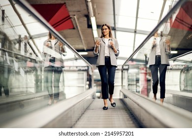 A Busy, Young Businesswoman Is Going Down The Escalator With Takeaway Coffee And Phone In Her Hands. A Woman Is Replying On Messages From Her Employees. A Businesswoman On The Escalator With A Phone