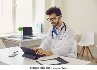 Busy young doctor using computer. Serious Caucasian male physician in medical lab coat and glasses sitting at office desk and working with electronic documents on modern digital tablet with keyboard