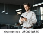 Busy young European business man using cellphone at work standing in office. Smiling male executive, businessman employee or entrepreneur holding smartphone working on mobile cell phone at work.