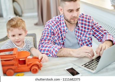 Busy Young Freelance Worker Wrapped Up In Finishing Project With Help Of Laptop While His Cute Little Son Playing With Toy Car, Living Room Interior On Background