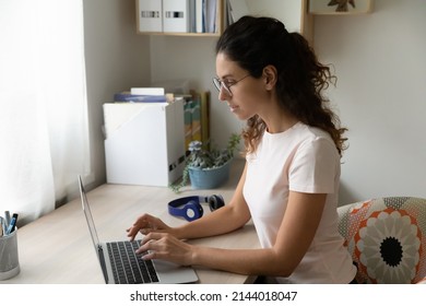 Busy young Hispanic business woman sit at desk wear glasses work on laptop texting e-mail solve business from homeoffice looking serious and confident, make telecommuting remote job from home concept