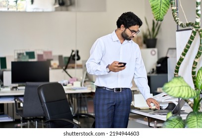 Busy Young Indian Business Man, Male Employee, Professional Manager Working On Laptop Holding Phone Standing In Modern Office Space At Work Using Corporate Software Technology Analyzing Market.