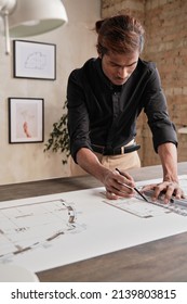 Busy Young Indian Man In Black Shirt Standing At Table And Using Ruler While Drafting House Plan In Loft Office With Minimalistic Pictures On Wall