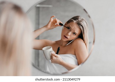 Busy young lady talking on smartphone, applying mascara and drinking coffee simultaneously in a hurry, being late for work, standing near mirror in bathroom