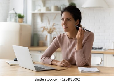 Busy young latin woman sit at home office by pc contact client using phone loudspeaker think on business offer idea. Hispanic female student search data at web via digital assistant listen to result