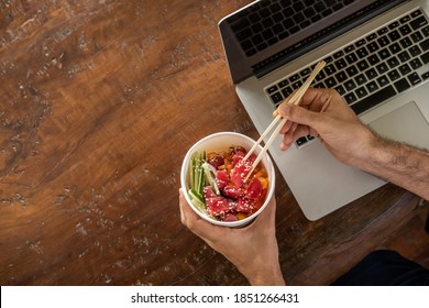 Busy Young Man In Casual Wear Sitting At Desk With A Laptop At Home Or In The Office And Eating Healthy Takeaway Poke Bowl Food From Container During Lunch Break. Healthy Lifestyle Concept. Top View