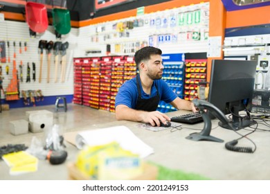 Busy young man employee working as a cashier at the hardware shop and using the computer to find a product or checking the inventory