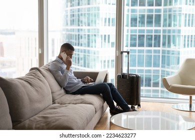 Busy young millennial travelling business man using digital gadgets for communication in hotel room, typing on laptop computer, talking on mobile phone, sitting on sofa at suitcase - Powered by Shutterstock