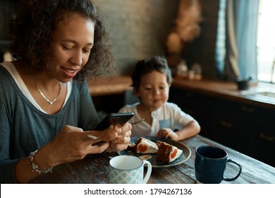 Busy Young Single Mother Using Cell Phone For Distant Work, Writing Posts On Social Network Accounts, Earning Money From Home On Self Isolation, Little Son Sitting At Table, Eating Cake In Background