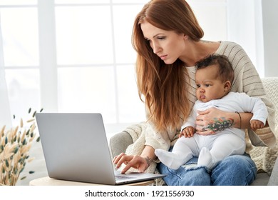 Busy young single parent caucasian mother using laptop computer distance working online from home office having video call virtual meeting with little cute african american infant baby girl daughter.