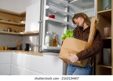 Busy Young Woman With A Heavy Bag Of Groceries Near The Refrigerator Talking On The Phone