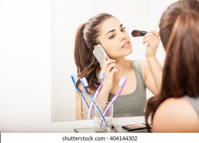 Busy Young Woman Taking A Call On Her Smartphone While Getting Ready For Work And Putting Some Makeup On