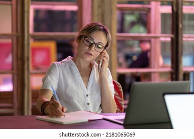 Busy Young Woman Talking On Smartphone, Making Notes While Studying In Public Library With Laptop. Focused Freelance Woman Preparing Her Project In Coworking Space. Education And Technologies Concept