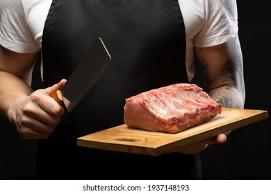 A Butcher In A Black Apron Holds Pork Ribs And A Meat Hatchet. Close-up.