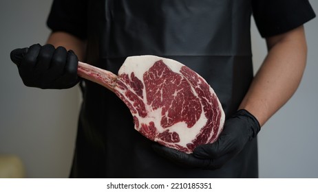 A Butcher With Black Apron And Black Rubber Gloves Is Holding Tomahawk MB5+ Steak With Hands