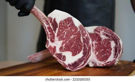 A Butcher With Black Apron And Black Rubber Gloves Is Holding Tomahawk MB5+ Steak With Hands