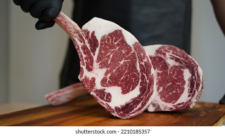 A Butcher With Black Apron And Black Rubber Gloves Is Holding Tomahawk MB5+ Steak With Hands