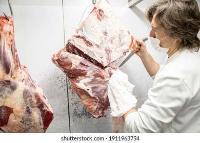 A Butcher Cutting A Large Piece Of Beef In A Refrigerated Room Of The Butcher's Shop. The Butcher Wears A White Uniform And Face Mask.