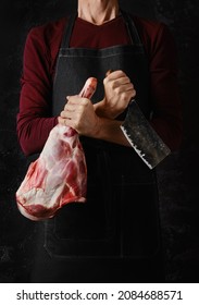 Butcher holds raw lamb leg in one hand and cleaver in another over dark background