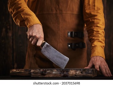 The butcher takes a large cleaver from the pocket of his leather apron.