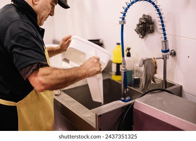 butcher wearing an apron washing trays and utensils in the sink of the butcher shop, carrying out cleaning and maintenance tasks in the establishment - Powered by Shutterstock