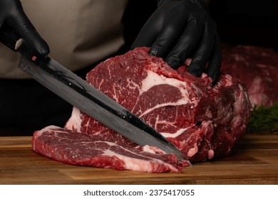 The butcher wearing black gloves cuts the meat with knife on wooden cutting board.Close-up piece of raw meat with a chef's knife on a dark background. - Powered by Shutterstock