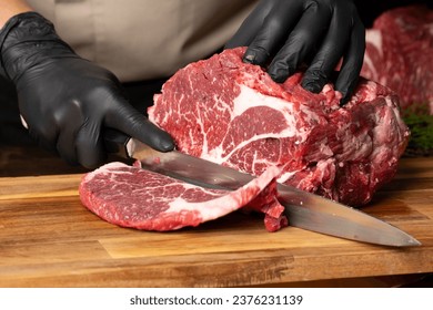 The butcher wearing black gloves cuts the meat with knife on wooden cutting board.Close-up piece of raw meat with a chef's knife on a dark background. - Powered by Shutterstock