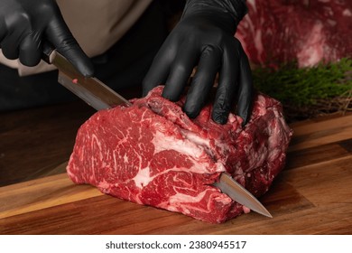 The butcher wearing black gloves cuts the meat with knife on wooden cutting board.Close-up piece of raw meat with a chef's knife on a dark background.