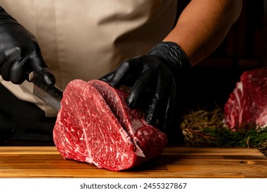 The butcher wearing black gloves cuts the meat with knife on wooden cutting board.Close-up piece of raw meat with a chef's knife on a dark background. - Powered by Shutterstock