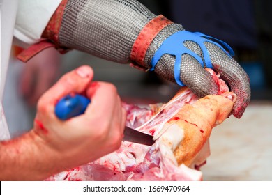 A butcher wearing protective gloves and using a sharp metal tool to cut and remove bone from a piece of pork in a busy butchery industry.