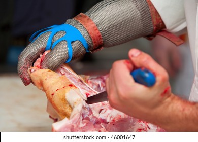 A butcher wearing protective gloves and using a sharp metal tool to cut and remove bone from a piece of pork in a busy butchery industry.