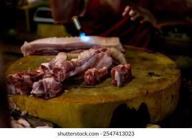 At a butcher's stall, a large piece of pork is being cleaned with a torch in the background, while another piece sits on a wooden cutting board in the foreground. Selective focus photo - Powered by Shutterstock