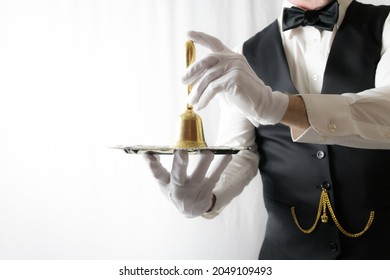 Butler Or Waiter In Black Vest And White Gloves Holding Gold Bell On Silver Serving Tray. Concept Of Service Industry And Professional Hospitality.