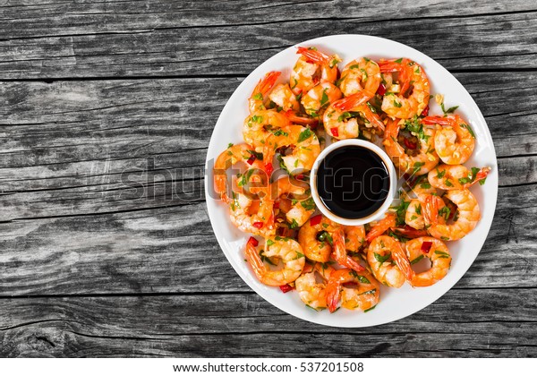 butter garlic fried shrimps
sprinkled with pieces of chilli and parsley on white dish with soy
sauce in small bowl in centre, on wooden table,view from
above