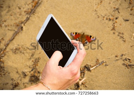 A butterfly in the sand is photographed by a girl on a white mobile phone on a sunny summer day.