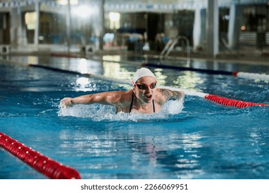 Butterfly swimming type. Young woman, professional female swimming athlete in cap and goggles training in pool indoors. Concept of sport, endurance, competition, energy, healthy lifestyle - Shutterstock ID 2266069951