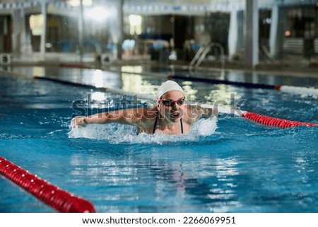 Butterfly swimming type. Young woman, professional female swimming athlete in cap and goggles training in pool indoors. Concept of sport, endurance, competition, energy, healthy lifestyle