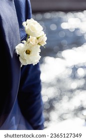 A buttonhole made of white flowers on a blue men's suit.Man's face and body are not visible.Wedding preparation and decoration in same style.Reflections of water are visible through blurred background