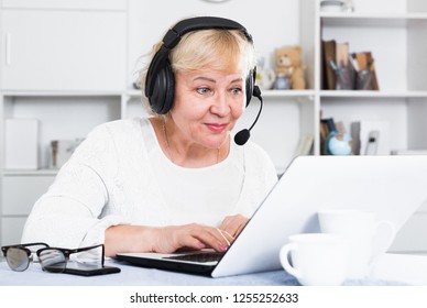 Buxom woman of an age sits in front of table with laptop in headphones with microphone