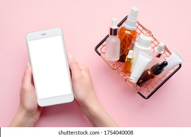 Buy Cosmetics Online Concept. Female Hands Holding Mobile Phone With Blank Screen Mockup And Shopping Basket Full Of Cosmetic Products On Pink Background. Flat Lay, Top View.