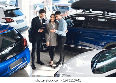 Buy Or Rent Car. Handsome Auto Vendor With Clipboard Talking To Couple, Undersign Contract