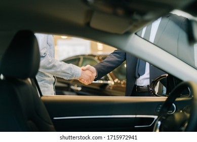 Buyer of car shaking hands with seller in auto dealership, view from interior of car. Close-up of handshake of business people. Concept of choosing and buying new car at showroom.