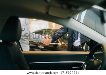 Buyer of car shaking hands with seller in auto dealership, view from interior of car. Close-up of handshake of business people. Concept of choosing and buying new car at showroom.