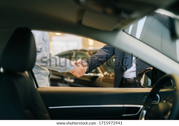 Buyer of car\
shaking hands with seller in auto dealership, view from interior of\
car. Close-up of handshake of business people. Concept of choosing\
and buying new car at\
showroom.